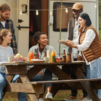 Young people smiling and chatting around picnic table filled with food and drinks for 4th of July BBQ. They are in front of an RV with lights strung overhead.